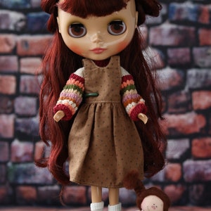 May include: A red-haired doll wearing a brown dress with a white and colorful crocheted sweater. The doll is holding a small, white and brown doll with a crocheted dress and long brown hair.