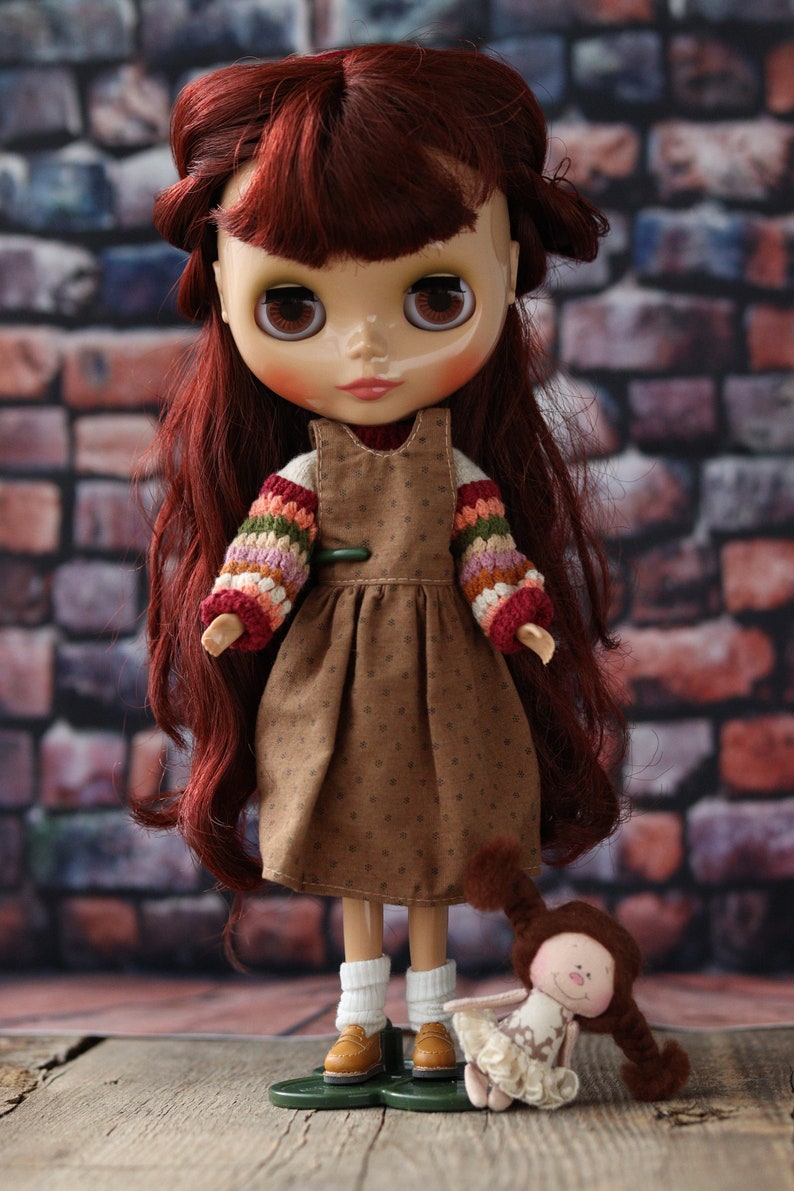 May include: A red-haired doll wearing a brown dress with a white and colorful crocheted sweater. The doll is holding a small, white and brown doll with a crocheted dress and long brown hair.
