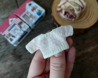 May include: A miniature, hand-knitted sweater in white with pink horizontal stripes. The tiny garment is held in a hand, with two small, pink and white boxes in the background. A dried rose and a wooden slice are also visible.