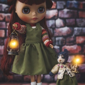 May include: A doll with long red hair wearing a green dress and holding a lantern. A small stuffed animal wearing glasses and a green dress is standing next to the doll.