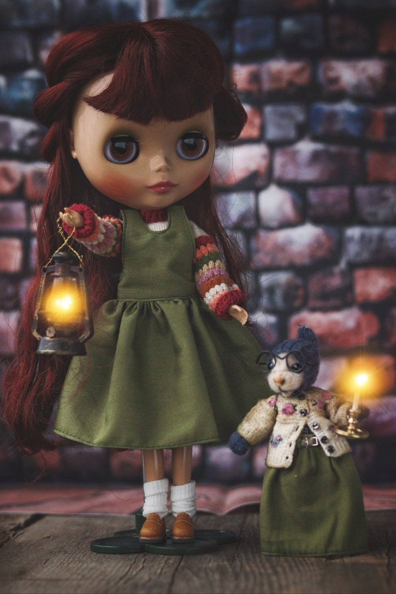 May include: A doll with long red hair wearing a green dress and holding a lantern. A small stuffed animal wearing glasses and a green dress is standing next to the doll.