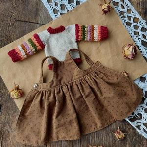 May include: A brown corduroy dress with a white button and a white and multi-colored striped sweater with red accents.