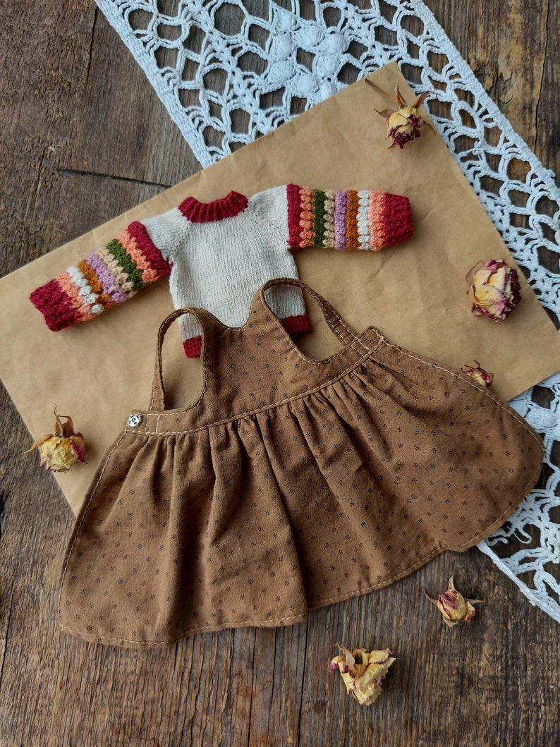 May include: A brown corduroy dress with a white button and a white and multi-colored striped sweater with red accents.