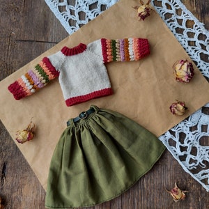 May include: A knitted sweater with colorful stripes on the sleeves and a green skirt with a brown belt. The sweater is cream colored with red trim. The skirt is a dark green.