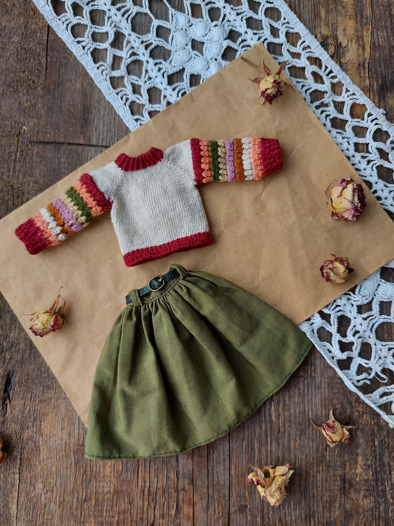 May include: A knitted sweater with colorful stripes on the sleeves and a green skirt with a brown belt. The sweater is cream colored with red trim. The skirt is a dark green.