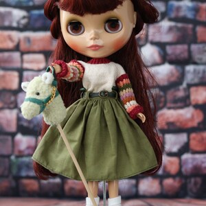 May include: A doll with long, auburn hair wearing a white sweater with colorful sleeves, a green skirt, and brown shoes. The doll is holding a stuffed horse on a stick.