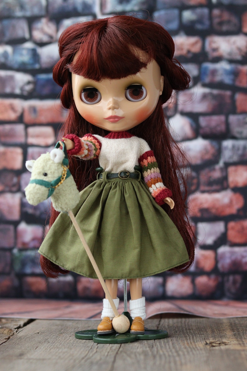 May include: A doll with long, auburn hair wearing a white sweater with colorful sleeves, a green skirt, and brown shoes. The doll is holding a stuffed horse on a stick.