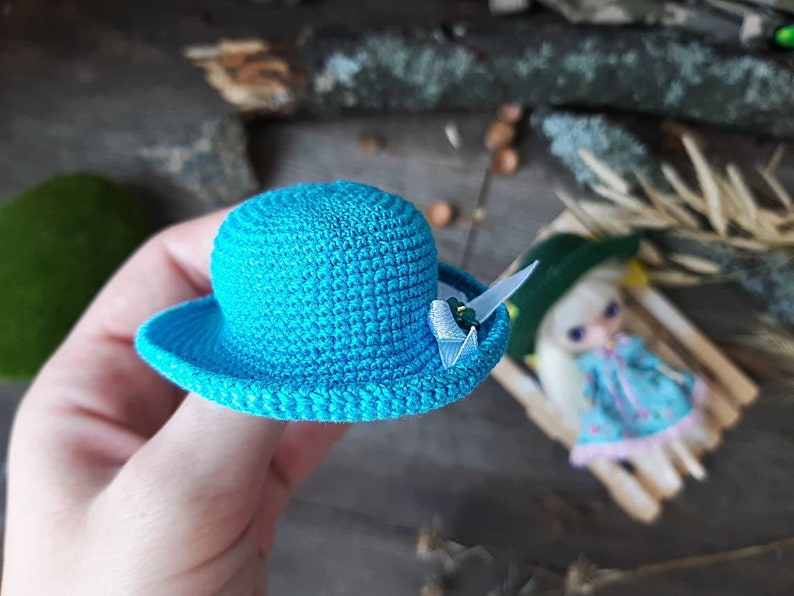 Pu&ograve; includere: Un piccolo cappello blu all'uncinetto con un nastro bianco e un accento di perla. Il cappello &egrave; tenuto da una mano.