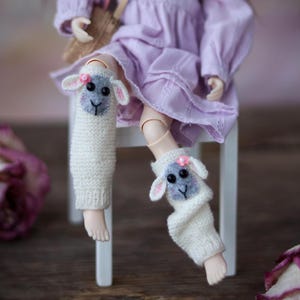 Può includere: Una bambola con un vestito lavanda e scaldagambe di lana bianca con un design a forma di viso di pecora. Gli scaldagambe hanno dettagli di fiori rosa.