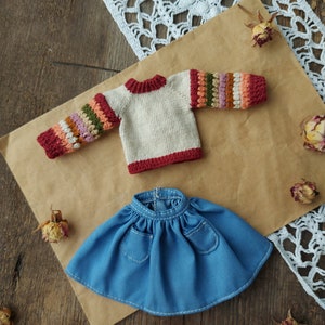 May include: A knitted doll sweater with a cream base and colorful stripes on the sleeves. The sweater has a red trim around the neckline and cuffs. A blue denim skirt with two pockets is also included.