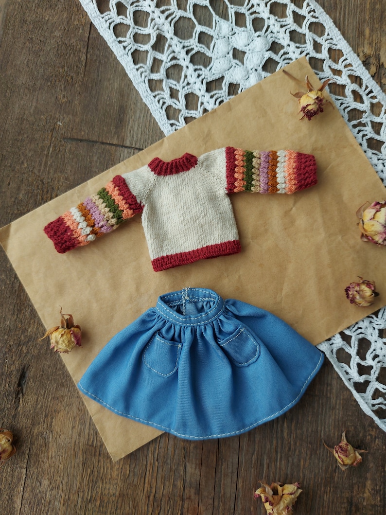 May include: A knitted doll sweater with a cream base and colorful stripes on the sleeves. The sweater has a red trim around the neckline and cuffs. A blue denim skirt with two pockets is also included.