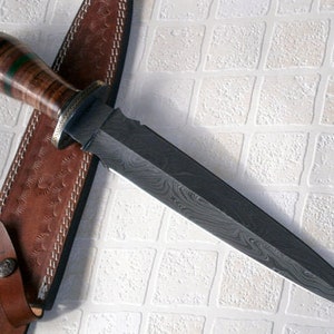 May include: A brown leather sheath with a decorative pattern holds a black and silver dagger with a wood handle. The dagger has a Damascus steel blade with a wavy pattern.