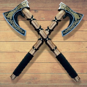 May include: Two crossed Viking axes with ornate wood handles and black leather wrappings. The axes have gold and black designs on the blades.
