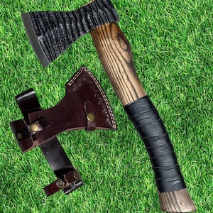 May include: A hand axe with a textured black metal head and a wooden handle wrapped in black leather. The axe includes a dark brown leather sheath with brass-colored snap buttons, designed to cover the blade and secure the handle for storage.