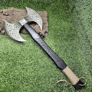 May include: A black-handled axe with a double-headed blade. The blade is decorated with a Celtic knot design and a skull. The axe is wrapped in brown twine near the handle.