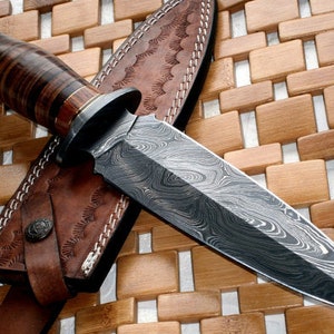 May include: A brown leather sheath with a decorative stitching pattern holds a silver dagger with a Damascus steel blade. The dagger has a brown wood handle with a decorative metal cap.