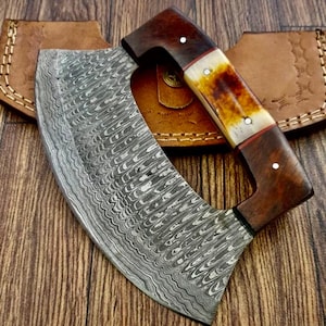 Op de afbeelding: Een gebogen, roestvrijstalen hakmes met een houten en beenen handvat. Het blad heeft een golvend damastpatroon. Het hakmes ligt op een houten oppervlak.