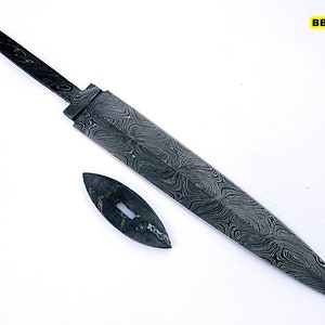 May include: A Damascus steel knife blade with a distinctive wavy pattern. The blade is approximately 12 inches long and has a pointed tip. A small, oval-shaped piece of metal with a hole in the center is also included.
