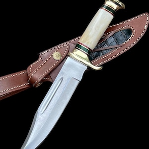 May include: A hunting knife with a polished steel blade and a bone handle, accented with brass and green details. The knife rests in a brown leather sheath with a black alligator-patterned insert. The sheath has a belt loop.