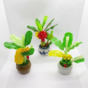 May include: Three miniature potted banana plants crafted from clay. Each plant features vibrant green and yellow leaves with clusters of small yellow, green, or red fruit. The plants are set in small, distinct ceramic pots in brown, white, and blue-patterned white.