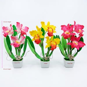 May include: Three artificial orchid plants in square white pots, each 8 inches in height. The left plant has 2-tone pink flowers, the center plant has yellow flowers with orange centers, and the right plant has ruby red flowers with green leaves.