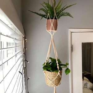 May include: Two plants hanging from a macrame plant hanger. The top plant is in a pink pot and the bottom plant is in a woven basket.