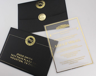 Pode incluir: Um convite de casamento em acrílico transparente com letras douradas. O convite apresenta os nomes do casal, "Aziza & Mikael", e um design decorativo. O convite é acompanhado por dois envelopes pretos com letras douradas e um selo dourado.