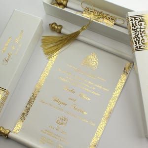 Puede incluir: Elegante pergamino de invitación de boda blanco con detalles dorados, presentado en una caja a juego. El pergamino presenta caligrafía dorada y bordes decorativos. La caja y el pergamino están adornados con intrincados patrones y texto dorados.