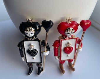 May include: Two enamel pin brooches featuring a playing card character with a crown and a heart-shaped balloon. One brooch is black and white, the other is red and white.