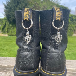 Puede incluir: Botas de cuero negro con costuras amarillas y un amuleto de pulpo unido a cada bota. Las botas tienen un patrón en relieve floral.