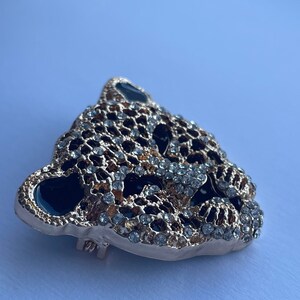 May include: Gold-tone brooch with a detailed design of a leopard's face, featuring black enamel and clear rhinestones.