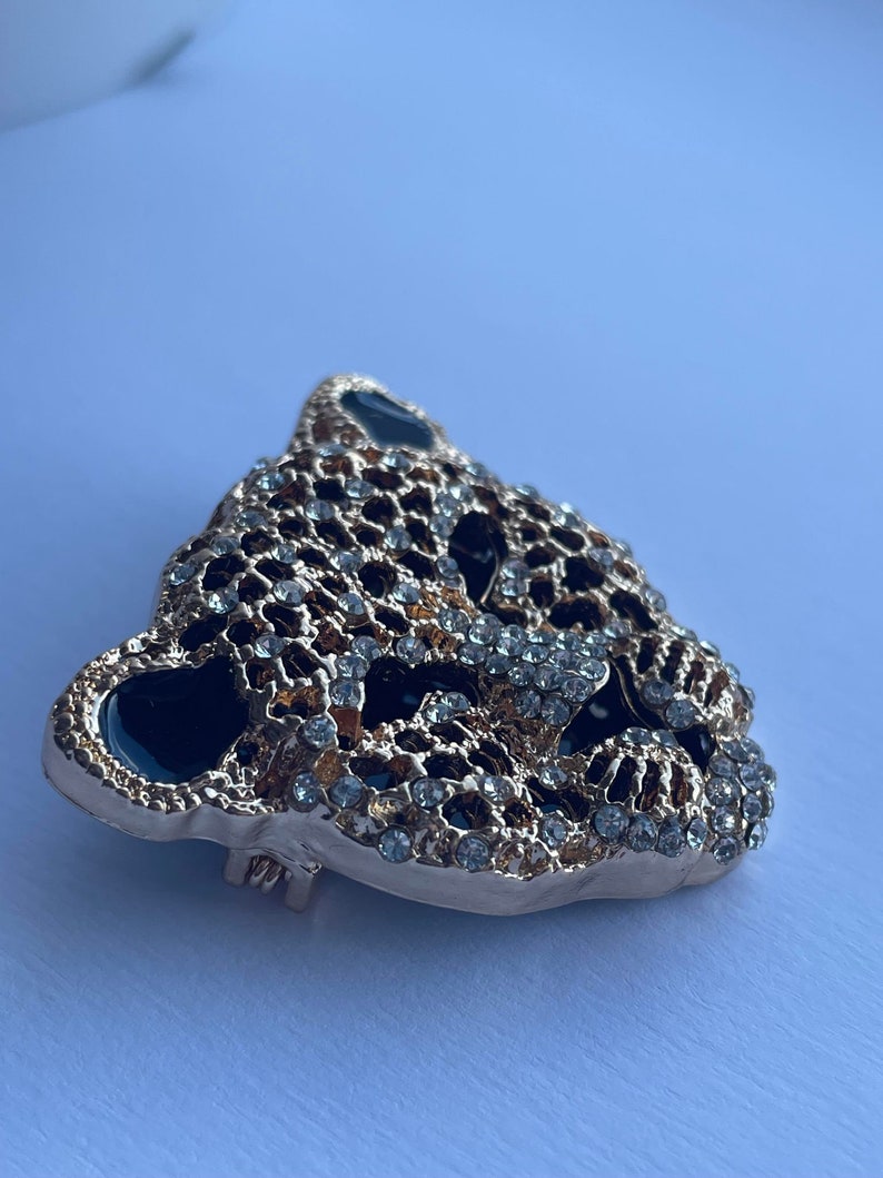 May include: Gold-tone brooch with a detailed design of a leopard's face, featuring black enamel and clear rhinestones.