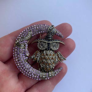 Pode incluir: Um broche dourado com uma lua crescente de cristal roxo e transparente com uma coruja usando um chap&eacute;u de bruxa preto em cima. A coruja &eacute; decorada com cristais laranja, transparentes e pretos.