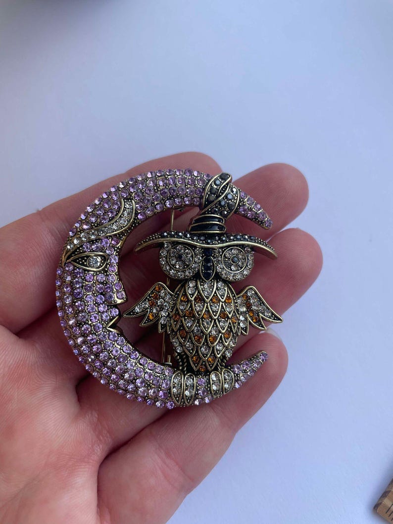 Pode incluir: Um broche dourado com uma lua crescente de cristal roxo e transparente com uma coruja usando um chap&eacute;u de bruxa preto em cima. A coruja &eacute; decorada com cristais laranja, transparentes e pretos.