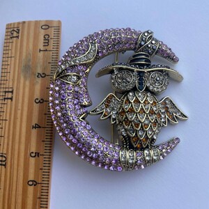 Pode incluir: Um broche dourado com uma lua crescente incrustada de cristais roxos e transparentes, com uma coruja usando um chap&eacute;u preto empoleirada no topo. A coruja tamb&eacute;m est&aacute; coberta de cristais.