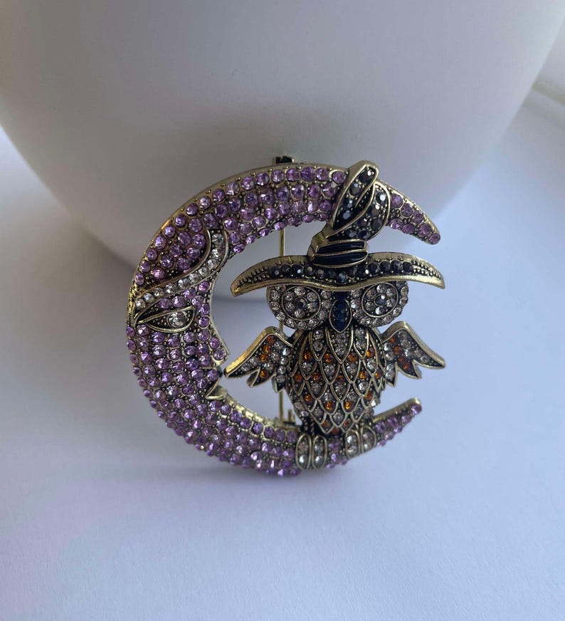 Pode incluir: Um broche dourado com uma lua crescente de cristal roxo com uma coruja usando um chap&eacute;u de bruxa. A coruja &eacute; decorada com cristais de v&aacute;rias cores, incluindo preto, branco, laranja e marrom.