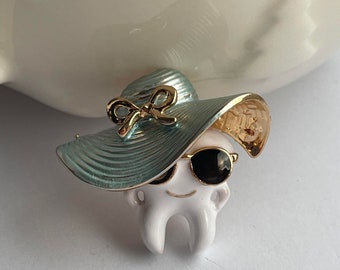 May include: A whimsical enamel brooch featuring a tooth character wearing a light blue hat with a gold bow and sunglasses. The tooth has a smiling face and is a fun accessory.