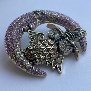 Pode incluir: Um broche dourado com uma coruja adornada com pedras preciosas pousada em uma lua crescente. A coruja &eacute; adornada com strass transparentes e &acirc;mbar, enquanto a lua &eacute; coberta com strass rosa.