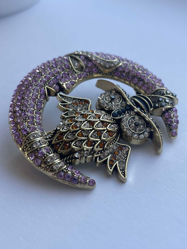 Pode incluir: Um broche dourado com uma coruja adornada com pedras preciosas pousada em uma lua crescente. A coruja &eacute; adornada com strass transparentes e &acirc;mbar, enquanto a lua &eacute; coberta com strass rosa.