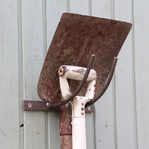 Puede incluir: Una pala de metal oxidada con un mango blanco y un gancho de metal unido al mango. La pala est&aacute; montada en una pared de madera.