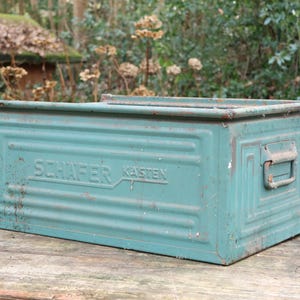 Puede incluir: Una caja de almacenamiento de metal turquesa con el texto "SCHAFER KASTEN" en el lateral. La caja rectangular tiene un asa y está colocada sobre una superficie de madera. La caja muestra signos de desgaste y mide unos 30 cm de largo.