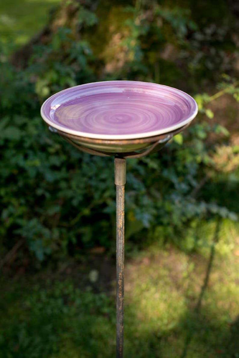 May include: A purple ceramic bird bath with a swirling pattern sits on a metal stand in a garden setting.