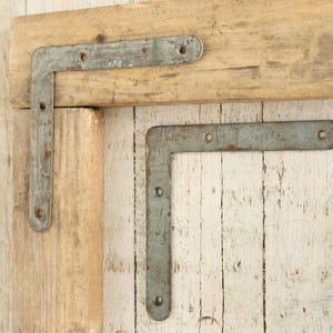 Puede incluir: Dos soportes de esquina de metal, uno fijado a una pieza de madera, el otro fijado a una superficie de madera pintada de blanco.