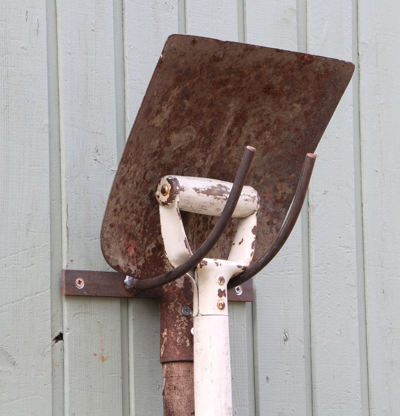 Puede incluir: Una pala de metal oxidada con un mango blanco y un gancho de metal unido al mango. La pala est&aacute; montada en una pared de madera.