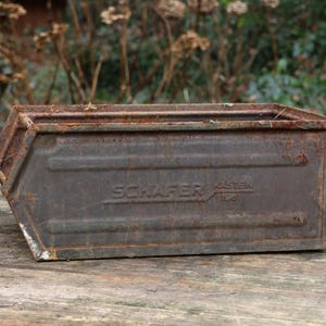 Könnte beinhalten: Ein rechteckiger, verwitterter Metallbehälter mit sichtbarem Rost und dem geprägten Text "SCHAFER". Der Behälter hat eine trapezförmige Form und steht auf einer Holzoberfläche, mit einem verschwommenen natürlichen Hintergrund.