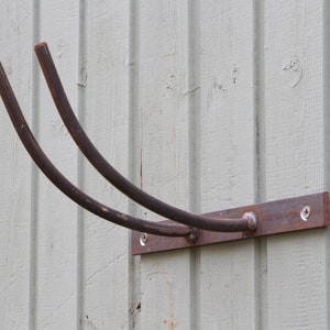 Puede incluir: Un gancho de pared de metal oxidado con dos brazos curvos. El gancho est&aacute; fijado a una pared de madera con tornillos.