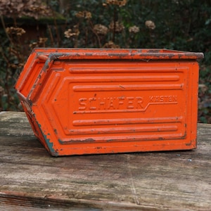 Puede incluir: Una caja de almacenamiento de metal naranja con el texto "SCHAFER KASTEN" en el lateral. La caja tiene forma rectangular con lados angulados y un aspecto ligeramente desgastado, sobre una superficie de madera.