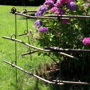 Puede incluir: Un soporte de planta de metal con tres ramas horizontales de madera, que sostiene un arbusto de hortensias rosas y moradas en flor. El soporte de la planta está en un área cubierta de hierba.