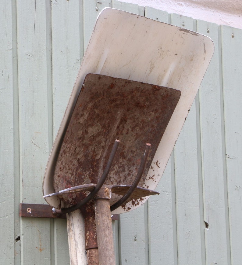 Puede incluir: Una pala de metal oxidada con un mango de metal blanco est&aacute; montada en una pared de madera. La pala est&aacute; colgada boca abajo con el mango apuntando hacia el suelo.