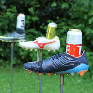 Könnte beinhalten: Drei Fußballschuhe auf Metallstangen, in denen jeweils eine Getränkedose steckt. Der vordere Schuh ist dunkelblau mit orangefarbenen Stollen, der mittlere ist weiß mit roten Akzenten und der hintere ist grau mit grünen Details. Jedes Paar enthält eine Dose.